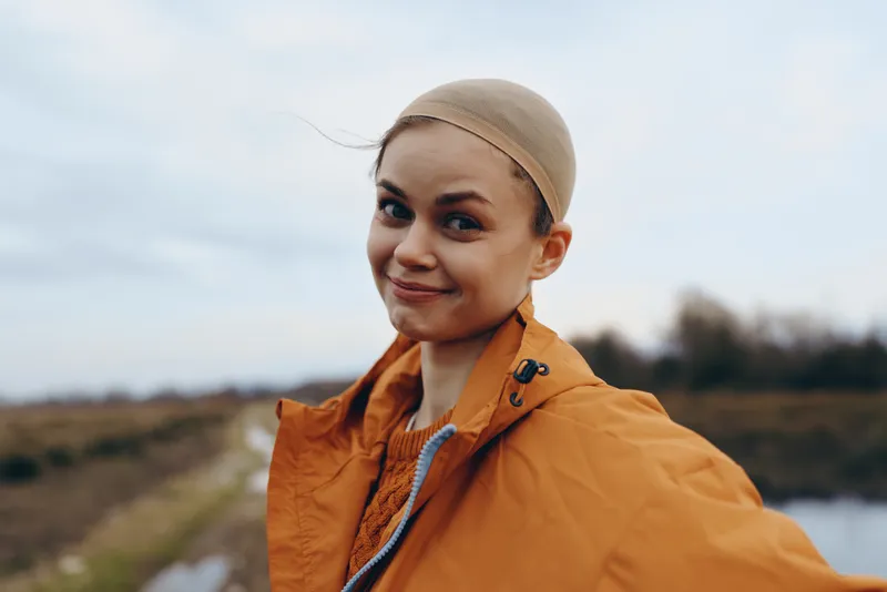 Woman lifestyle outdoor portrait outdoors countryside smiling, orange jacket, scarf, headscarf, gentle expression, countryside field and road blurred background, natural light, calm travel mood