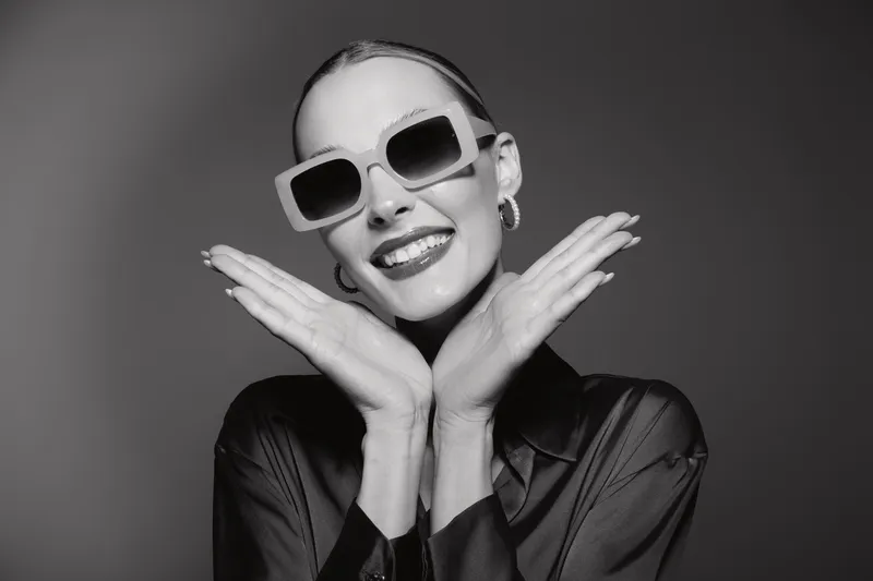 Stylish young white woman with light skin wearing dark sunglasses and black satin blouse smiles confidently on grey background. Concept of fashion, elegance, and self-expression.