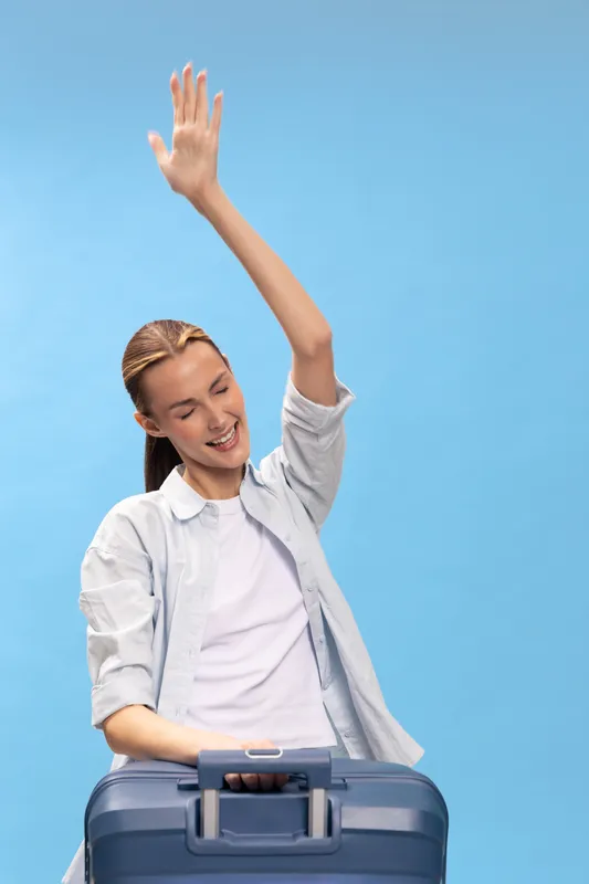 Happy young woman with suitcase raises hand and smiles, wearing casual clothes on blue background. Concept of travel, vacation, journey and positive emotions.