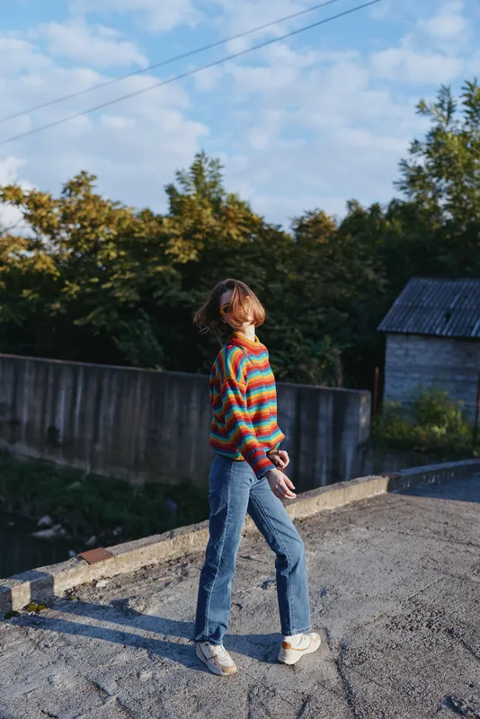 Young woman in her early 20s wearing a colorful striped sweater and blue jeans, walking outdoors on sunlit concrete by a canal, short hair, casual outfit, relaxed lifestyle travel portrait.