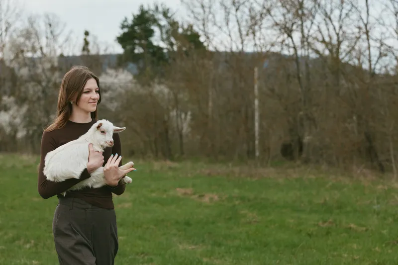 Woman outdoors holds a white goat kid in her arms, enjoying a calm moment in a grassy field, showcasing rural lifestyle and animal care, serene mood, natural light, authentic countryside scene
