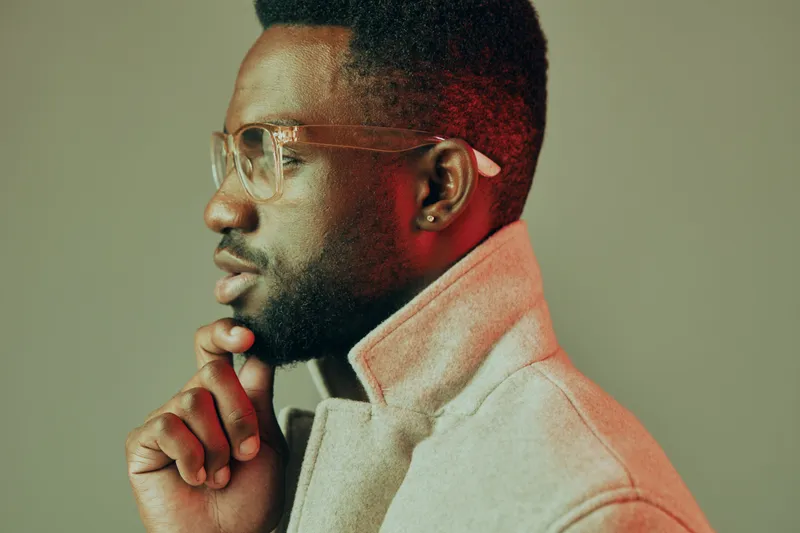 Stylish African man in glasses contemplating thoughtfully, wearing a fashionable beige coat against a colorful gray background, showcasing modern elegance and confidence