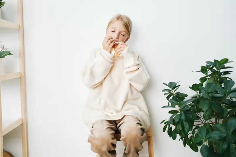 A person sits on a small wooden stool wearing a fluffy cream sweater, savoring a snack in a bright, minimal room with soft shadows. The scene exudes comfort and warmth, emphasizing texture and quiet,