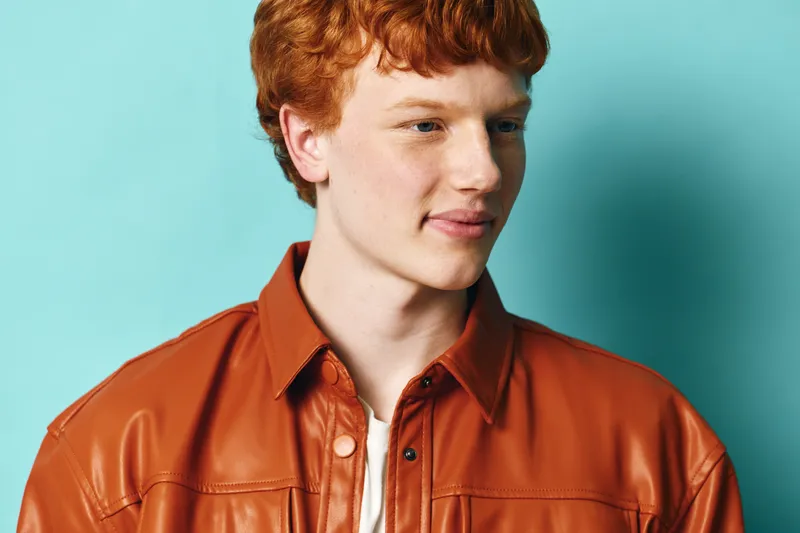 Young man with red hair wearing a brown leather jacket standing against a blue background. Portrait with casual style and natural expression looking to the side.