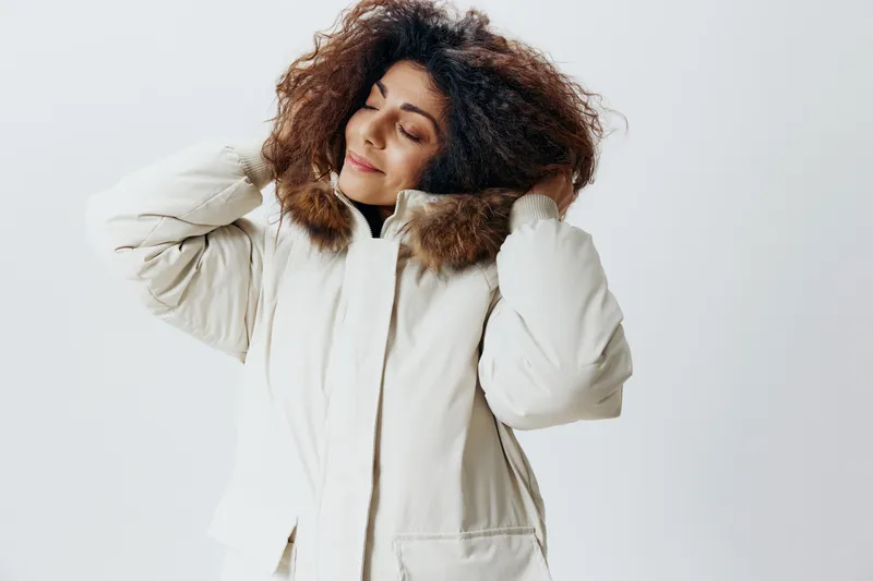 Веселая кудрявая симпатичная латиноамериканка в капюшоне из меха мягкого теплого бежевого зимнего пальто позирует изолированно с закрытыми глазами на белом фоне студии