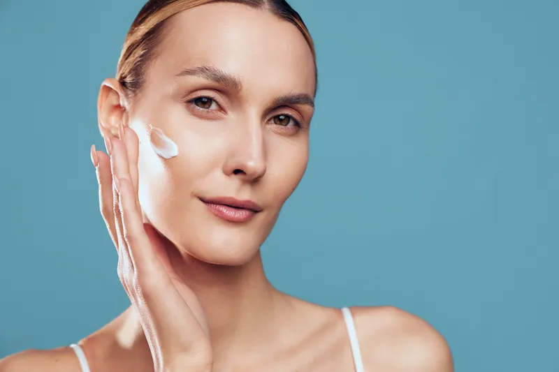 Young woman applying moisturizer on face, clear skin, gentle smile, skincare routine, soft natural light, blue background, beauty concept, closeup portrait