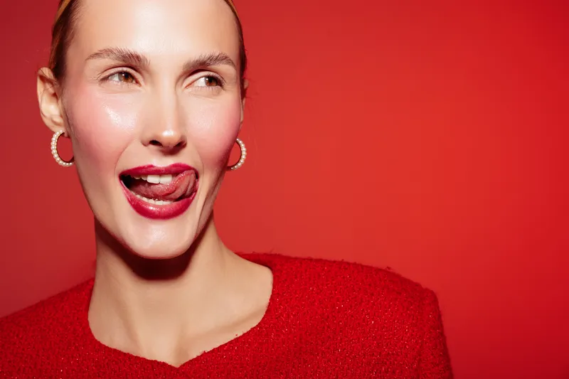 Young woman with red lipstick and earrings smiles playfully, licking her lips against a red background. Concept of beauty, fashion, and confident female expression in studio lighting.