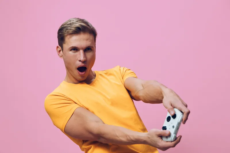 surprised young man playing video game holding white gamepad isolated on pink background digital entertainment concept