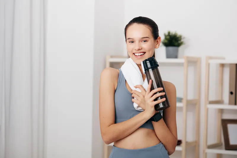 Fitness woman in athletic wear holds a water bottle and towel, smiling in a bright home gym. Clean background with soft light conveys health, wellness, and active training in a modern space.