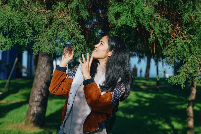 Woman enjoying outdoor water spray under evergreen trees in a sunny park, casual jacket and scarf, joyful moment amid nature, fresh air and playful refreshment, carefree summer vibe