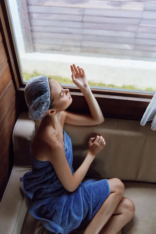 Relaxed woman wrapped in blue towel and shower cap sits by window in cozy wooden bathroom, enjoying peaceful moment and soft natural light indoors.