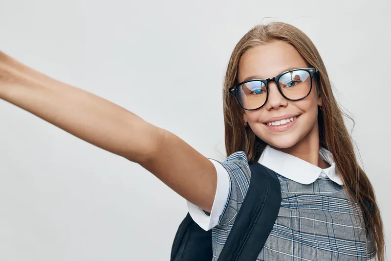 Girl smiling with glasses and backpack posing for selfie with outstretched arm in casual outfit on gray background. Young student happy and confident with long hair.