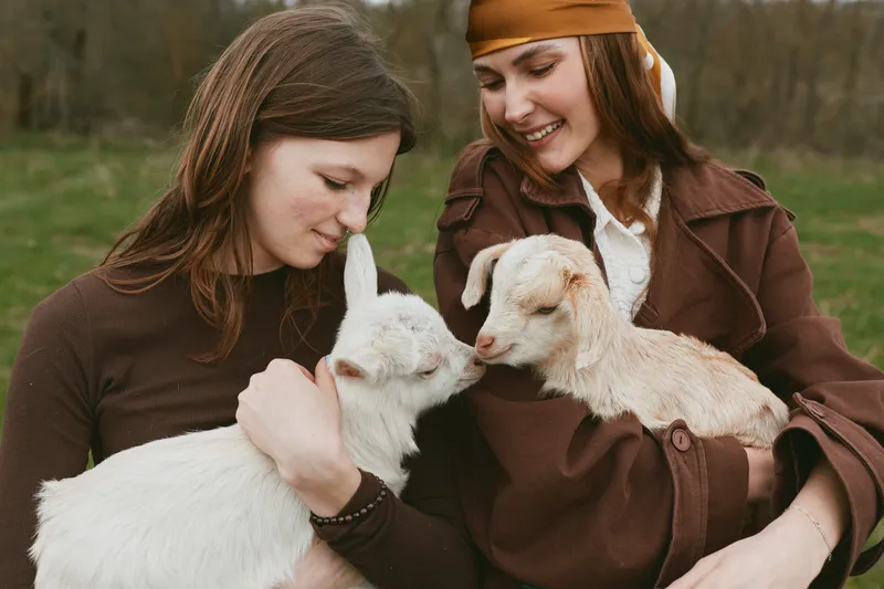 Two women in brown jackets share a tender moment with calm goats in an open field, a bright rural lifestyle scene emphasizing companionship, gentle care, and simple pastoral joy in nature.