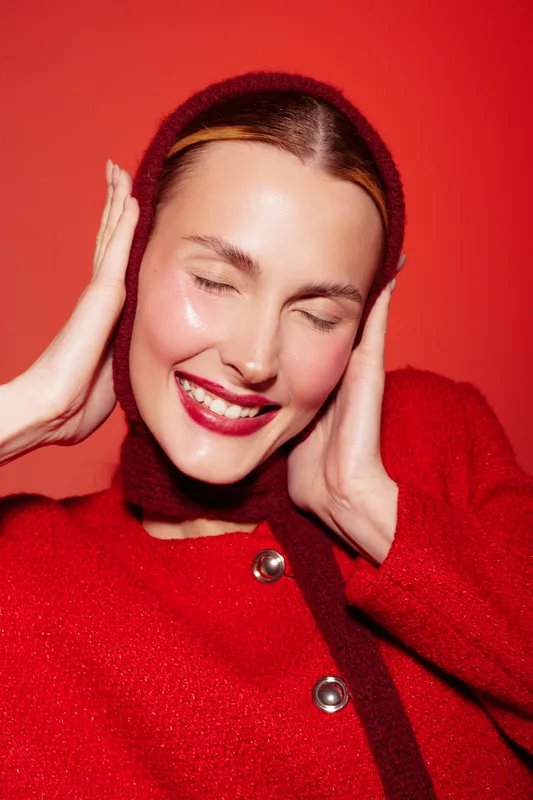 Smiling woman in red sweater and headscarf poses with closed eyes and bright lipstick on red background. Concept of happiness, fashion, and beauty in studio portrait.