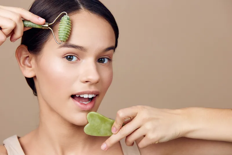 Young woman using jade roller and gua sha tool on her face for a skincare routine, showcasing natural beauty and wellness practices