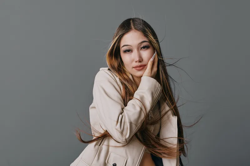 Young woman with long hair and casual beige jacket posing with thoughtful expression against grey background. Beauty, fashion, and portrait photography with natural makeup and hairstyle.
