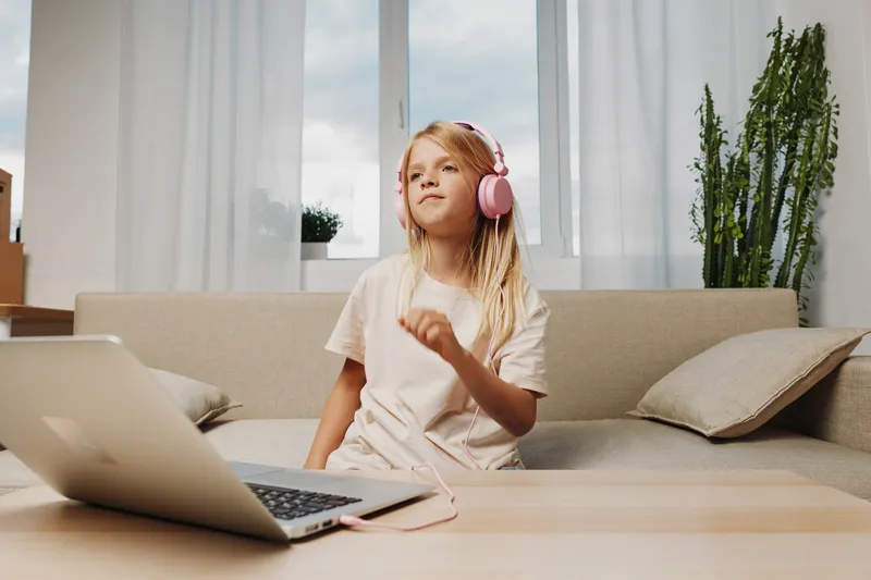 Blond girl with pink headphones listens to music and enjoys dancing at home in bright living room with laptop. Concept of childhood, leisure, technology, and happiness.