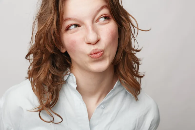 Young woman with curly hair wearing white shirt looks sideways with playful expression on light background. Concept of casual style, youth, and natural beauty portrait.