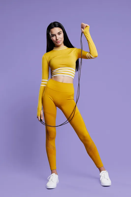 Sport fitness woman in bright yellow activewear holding a skipping rope, standing against a purple background. Healthy lifestyle, energy, athletic pose.