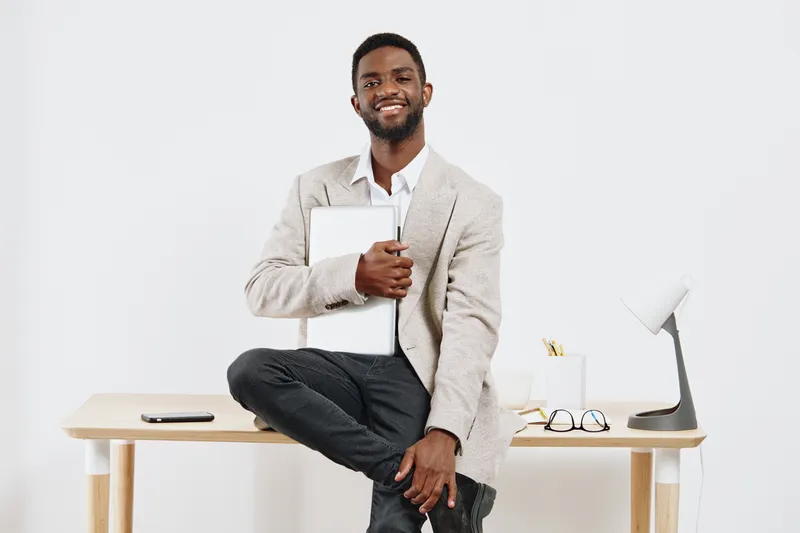 Confident young smiling man with dark skin, wearing formal blazer and white shirt, holding laptop, standing in modern office environment with minimalistic white background, professional business peopl