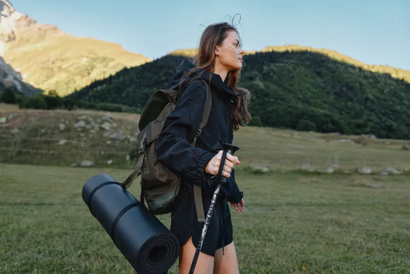 A solitary hiker with a backpack and rolled mat walks through a green valley under clear skies, ready for adventure and outdoor discovery.