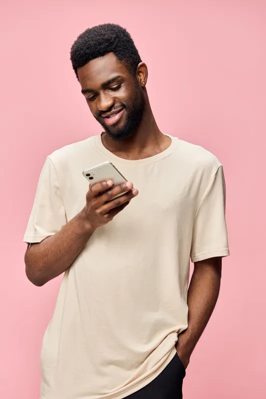 Smiling young African American man holding smartphone, wearing casual beige t shirt, against soft pink background, enjoying social media moments and connection