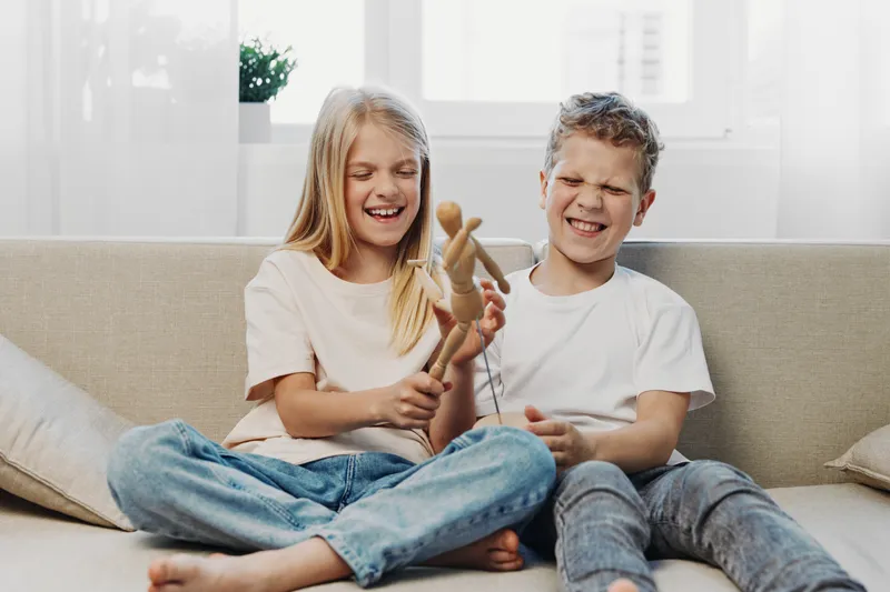 Two children playing with wooden puppet on couch, smiling and laughing in bright living room. Concept of childhood fun, sibling bonding, and creative playtime.