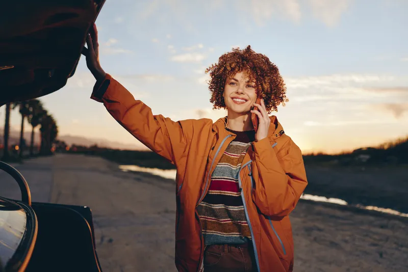 Woman outdoor lifestyle at sunset, smiling curly hair, wearing orange jacket, standing by car on road, one arm raised, warm light, relaxed travel mood