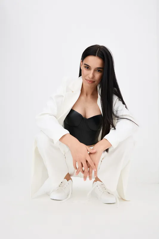 Young woman in stylish white suit and black corset crouching with relaxed pose, long dark hair, minimalist white background, modern fashion and casual style.