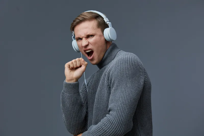Young caucasian man wearing headphones singing enjoying music in grey sweater isolated on grey background digital detox mental health concept