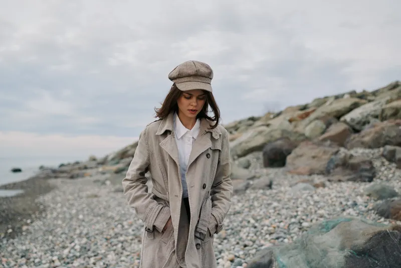 Young woman in beige coat and hat walks on rocky beach under cloudy sky. Casual fashion style with natural landscape. Concept of autumn, solitude, and outdoor lifestyle.