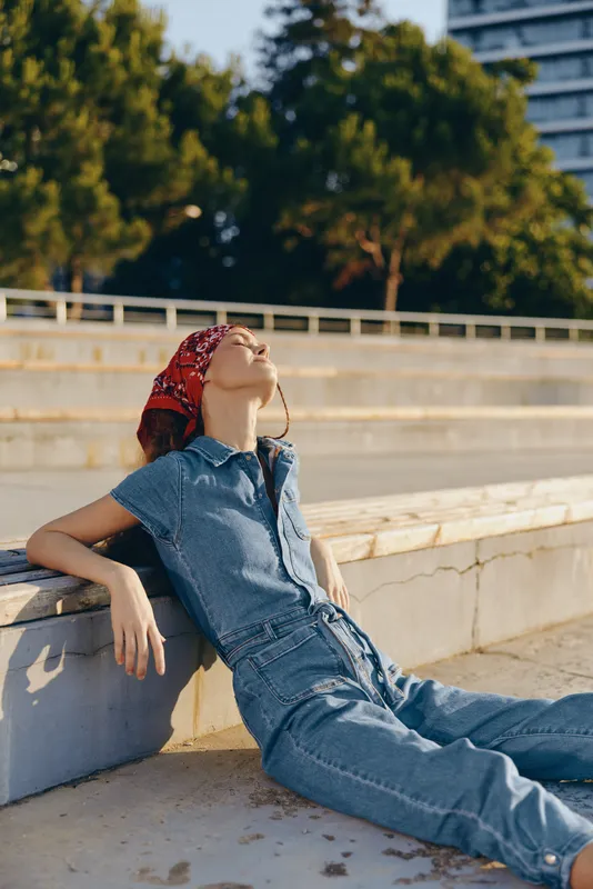 Lifestyle woman in modern boho-western denim street style relaxes outdoors with calm expression, casual outfit, vibrant film color capturing warm sunny atmosphere.