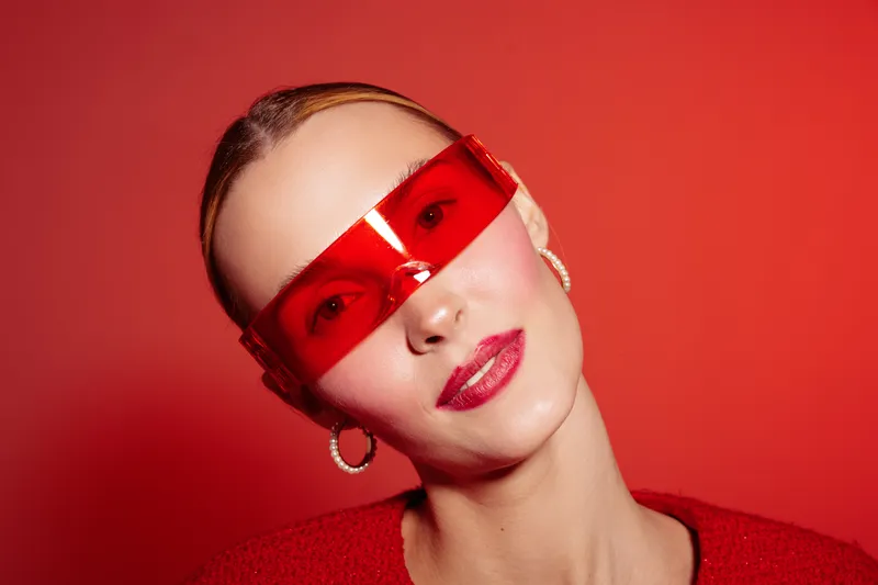 Portrait of a stylish woman wearing red transparent sunglasses and red lipstick against a red background. Concept of fashion, beauty, modern style, and bold accessories.
