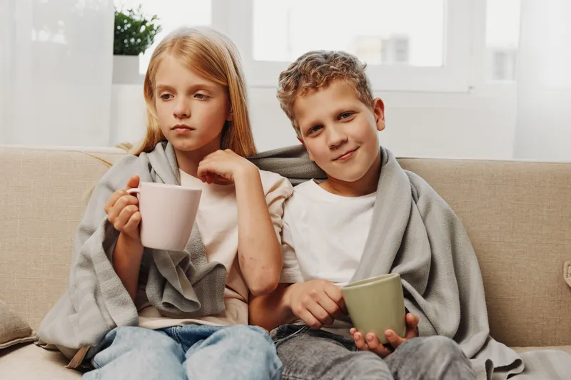 Two children wrapped in gray blankets sit on a beige sofa holding mugs. The bright room with window light creates a cozy, calm atmosphere of relaxation and comfort at home.