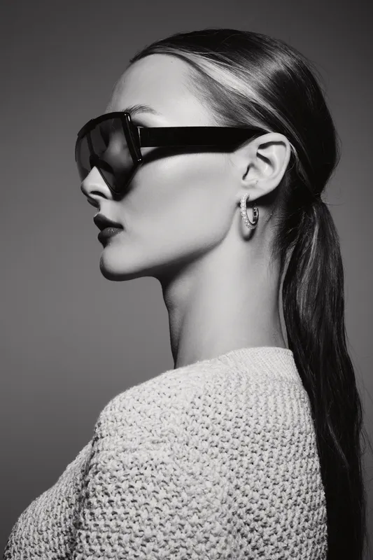 Profile of Caucasian woman with fair skin wearing sunglasses and textured sweater on grey background.