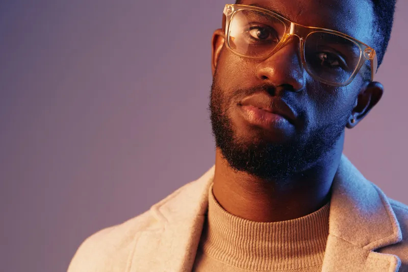 Stylish portrait of a young Black man in glasses wearing a light sweater, showcasing confidence and modern fashion against a colorful gradient background
