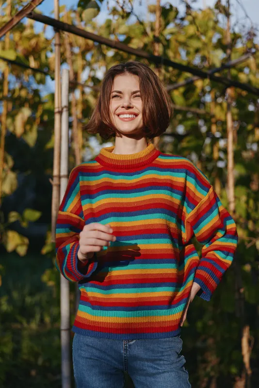 Woman smile colorful sweater outdoors portrait of a happy casual young woman in a striped knit pullover, standing in a sunlit vineyard with relaxed pose and cheerful expression.