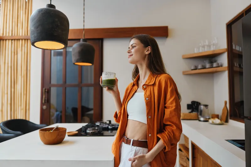 Весёлая молодая женщина наслаждается зелёным смузи на современной кухне