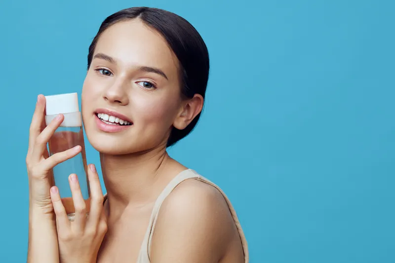 Young woman presenting a skincare product with a cheerful expression against a blue background, showcasing healthy skin and beauty