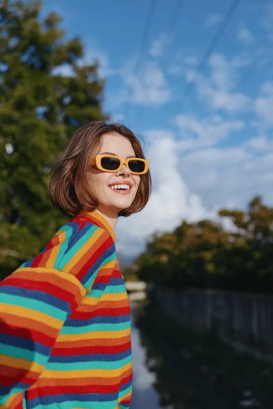 Woman smile rainbow sweater sunglasses portrait outdoor happy fashion model by river and trees, enjoying sunny day with colorful knit, youthful energy, confident pose and natural light.