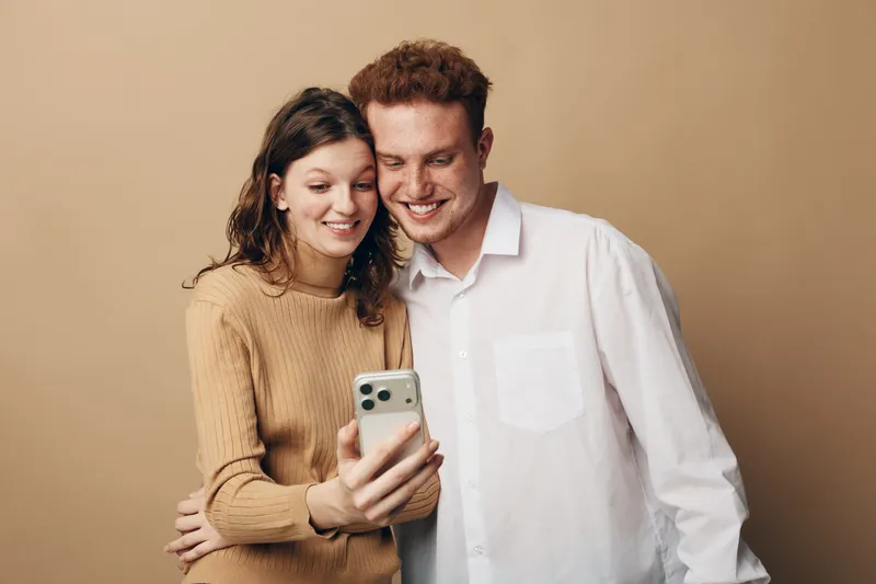 Young couple smiling and taking a selfie with smartphone on beige background. Casual clothing, happy mood, and modern technology concept for relationships and social media.