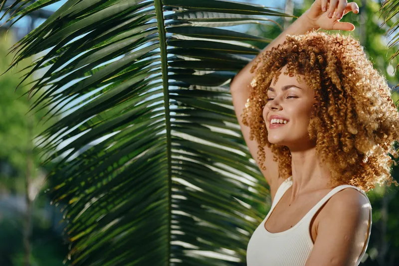 smiling woman with curly hair enjoying tropical sunlight beside large green palm leaf in summer outdoor setting, natural beauty and happiness captured.