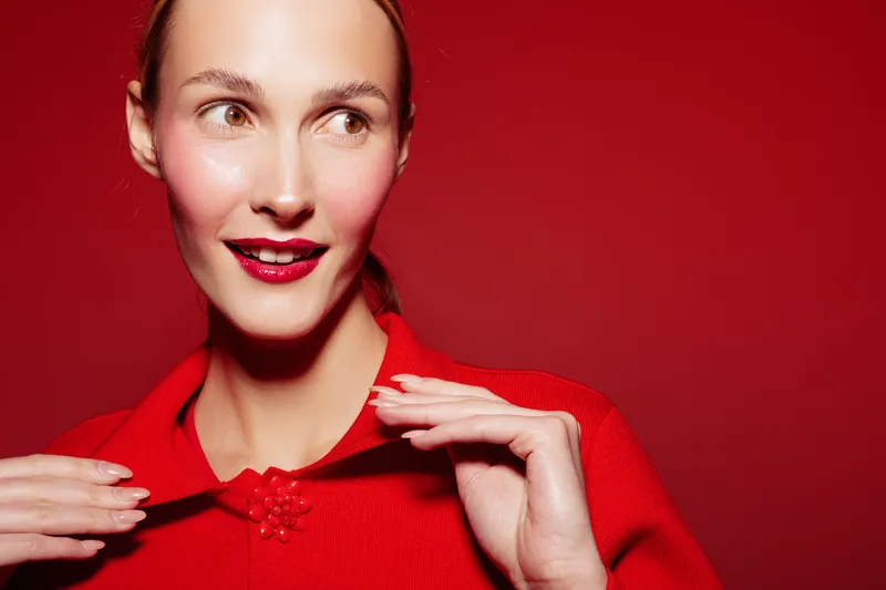 Young woman with red lipstick and red sweater looks aside with smile on red background. Close-up portrait with bright colors and confident mood. Concept of fashion, beauty, and style.