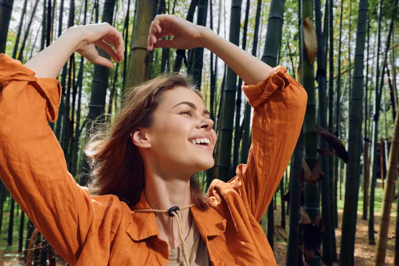 woman smile bamboo forest portrait joyful young female outdoors, candid closeup of a smiling woman in orange blouse with raised arms, sunlight and wind in hair, natural lifestyle freedom.