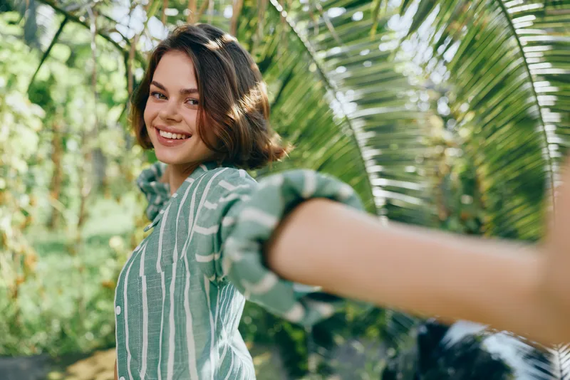 Bright outdoor portrait of a smiling woman in a lush garden, reaching toward the camera with an outstretched arm, framed by tropical leaves.