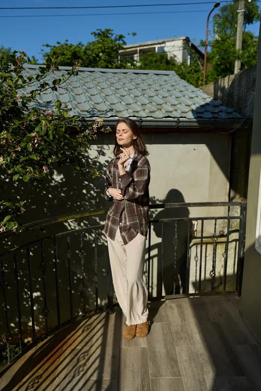Woman in plaid shirt and light trousers stands on a balcony, natural sunlight highlighting a casual, relaxed lifestyle and urban mood, with warm tones and a tranquil, everyday fashion feel