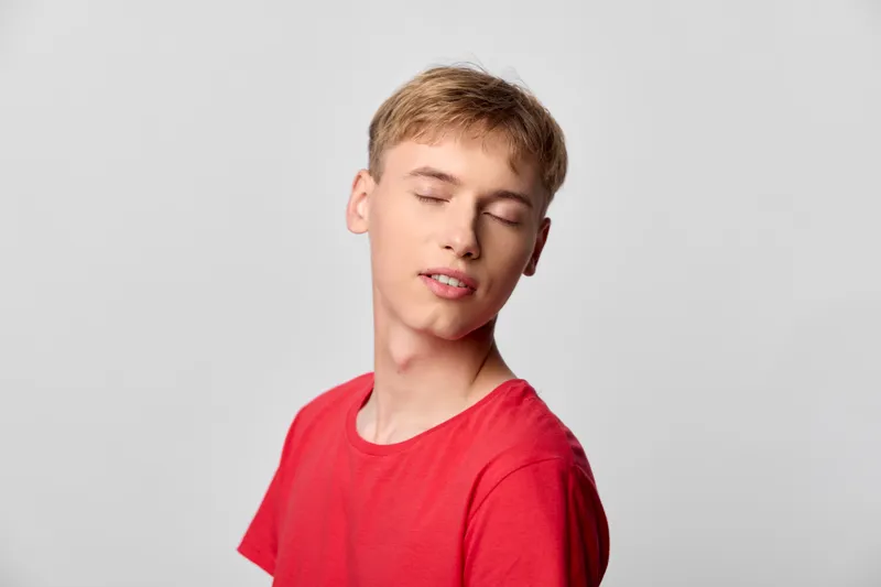 Close up of a young man with eyes closed, wearing a bright red shirt, in a minimal studio setting. The relaxed pose, soft lighting, and serene expression convey calm and quiet mood.
