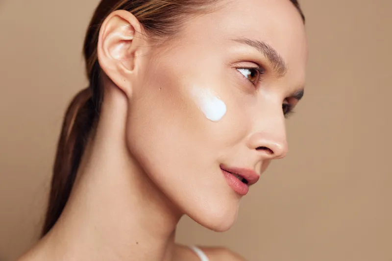 Closeup of young woman with natural skin applies facial cream on cheek looking sideways with calm expression isolated on beige background in skincare beauty concept.