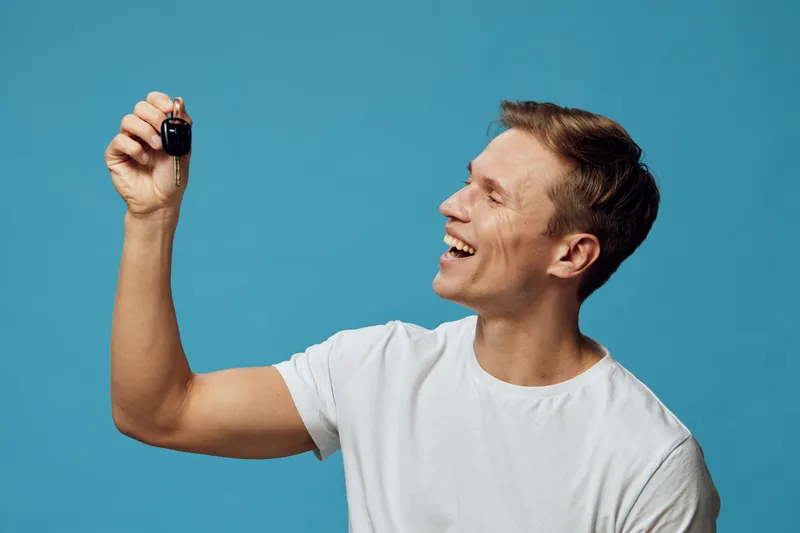 smiling young caucasian man wearing white t-shirt look at car key fob hold in hand isolated blue background cyber security concept