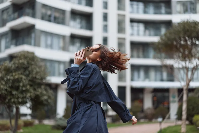 Young woman in a navy trench coat dances outdoors, wind flips her hair in a bright urban courtyard, capturing a joyful carefree moment against modern glass buildings.